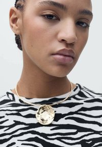 Gouden hangende ketting aan een dunne, gebogen metalen ketting; de hanger heeft een gestructureerde, organische vorm. Model draagt een zwart-wit gestreepte zebratop.