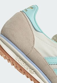Chaussure de sport avec un dessus en cuir suédé gris, des accents bleu clair, un tissu blanc et une semelle en caoutchouc pêche ; présente un logo en relief et des coutures texturées.