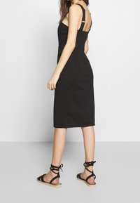 Kobieta w dopasowanej czarnej sukience midi z grubymi regulowanymi ramiączkami oraz czarnych płaskich sandałach na sznurówki, stojąca bokiem na jednolitym tle.