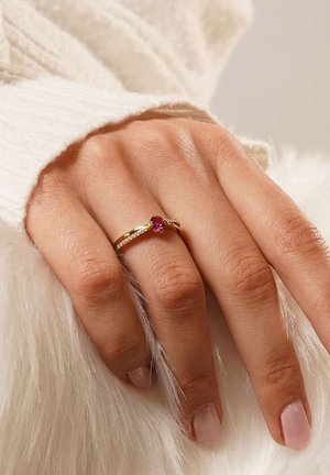 Goldring mit einem zentralen roten Edelstein, verziert mit kleinen klaren Steinen entlang des Bands. Die Hand ruht auf einer strukturierten weißen Felloberfläche.