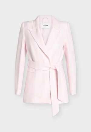 Kvinde med kort blond hår iført en lyserød blazer med bælte, brede cremefarvede bukser, holder en lille sølv håndtaske, står på hvid baggrund.