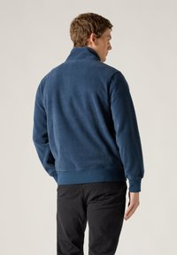 Μπλε μπουφάν φλις με ψηλό γιακά, ριμπ μανσέτες και ζώνη στη μέση. Απαλή υφή και απλός σχεδιασμός, εμφανίζεται σε άτομο από πίσω.