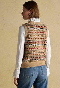 Beige kudum, millevärviliste lillemustertega. Kõrge kaelusega valge pluus all, teksased, millel on nähtavad taskud.