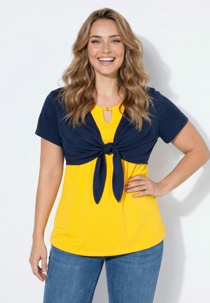 Lachende vrouw met golvend blond haar die een gele top en een gebonden marineblauwe kurzarm shrug draagt, poserend met één hand op haar heup.