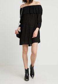 Schwarzes schulterfreies Kleid mit Rüschendetail, langen Ärmeln und lockerer Passform. Kombiniert mit schwarzen Stiefeletten mit metallischen Akzenten.