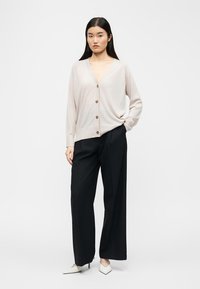 Femeie tânără purtând un cardigan bej deschis cu nasturi, pantaloni negri cu picioare largi și pantofi cu toc ascuțit albi, stând cu o mână în buzunar.