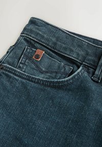 Mörkblå denimjeans med synlig sömnad, en liten ficka och ett läderdetalj med en metallknapp. Strukturerad tyg.