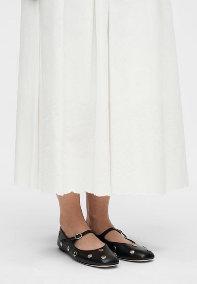 Γυναίκα που φοράει λευκή μίντι φούστα με κέντημα και μαύρα παπούτσια Mary Jane με ασημένιες λεπτομέρειες καρφιτσών σε λευκό φόντο.