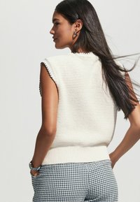 Krämfärgad, utan ärmar, stickad topp med ribbad nederkant och svarta vågiga detaljer, tillsammans med rutiga shorts; mjuk textur och avslappnad passform.