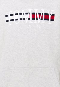 Světle šedá mikina s vyšitým nápisem "TOMMY JEANS" v bílé barvě, s černými a červenými horizontálními pruhy nad textem. Měkká textura, klokaní kapsa.