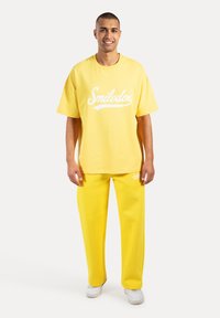 Gele katoenen oversized t-shirt met wit "Smilodox" logo, gecombineerd met bijpassende gele broek. Model staat voor een neutrale achtergrond.