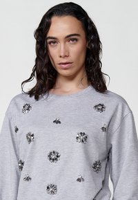 Felpa grigia con maniche lunghe, decorata con diverse embellishments in strass disposte in forme floreali sulla parte anteriore.
