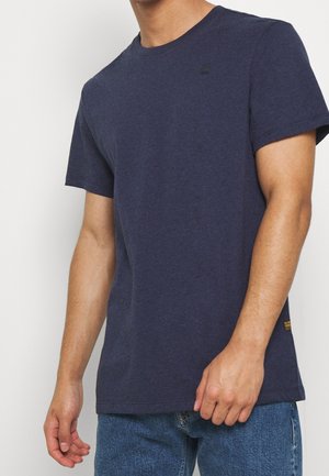 Man in een effen marineblauw T-shirt met korte mouwen en een blauwe spijkerbroek, van nek tot heupen, tegen een witte achtergrond.