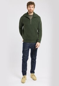 Un pull en tricot vert foncé avec un col zippé quart, porté sur une chemise grise, associé à un jean bleu foncé et des chaussures en daim beige clair.