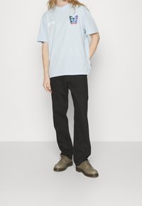 Light blue cotton t-shirt with a butterfly graphic on the chest, paired with black pants and brown shoes. Simple design, casual fit.