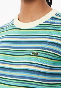 Stribet kortermet sweater i blå, grøn og gul med ribbet tekstur. Har en gråhvid rund halsudskæring og et lille krokodillelogo.
