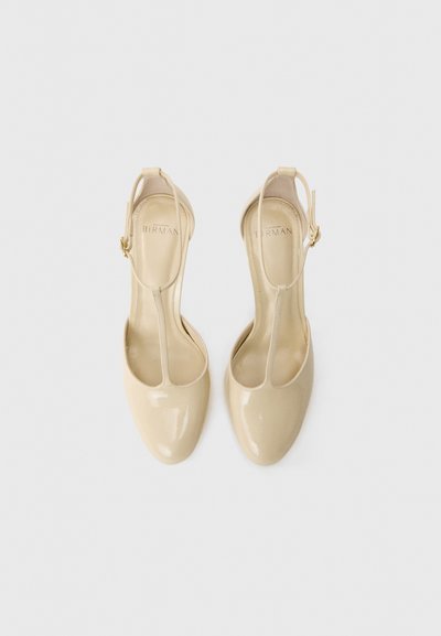 Beige patent leather high-heeled sandals with a T-strap design and ankle buckle. Smooth texture and pointed toe shape. Gold hardware accent.