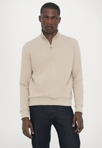 Beige kvartdraget sweatshirt med en slät textur och ribbade mudd, matchad med mörka jeans. Har en minimalistisk design.