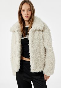 Hvid, fluffy faux fur jakke med krave og frontknaplukninger. Bæres over en sort cropped t-shirt med rund halsudskæring og korte ærmer.