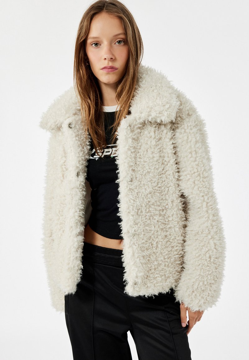 Hvid, fluffy faux fur jakke med krave og frontknaplukninger. Bæres over en sort cropped t-shirt med rund halsudskæring og korte ærmer.