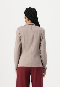 Kvinna med långt vridet hår, klädd i en taupefärgad kavaj och vinröda byxor, som står vänd mot en helt vit bakgrund.