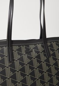 Čierna taška na rameno s textúrovaným povrchom a geometrickým zlatým vzorom. Obsahuje dve čierne uši a malú detailnú značku Lacoste.