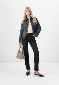 Woman wearing dark denim jacket and pants, beige top, black heels, holding beige snakeskin-patterned handbag, standing against white background.