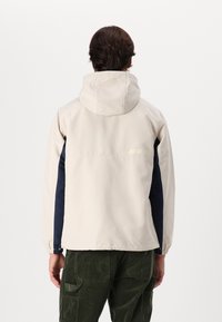Light beige hooded jacket with navy side panels, smooth texture, and subtle logo detail at the back. Features elastic cuffs and hem.