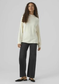 Cremefarbenes geripptes Sweatshirt mit hohem Kragen, kombiniert mit weit geschnittenen, dunklen Jeans und schwarzen Slippern. Glatte Textur und lockere Passform.