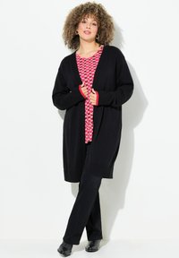 Zwarte gebreide cardigan met open voorkant en ribbels aan de randen, gedragen over een rood patroon t-shirt. Gepaard met zwarte broek en zwarte schoenen.