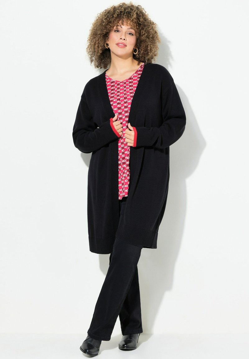 Zwarte gebreide cardigan met open voorkant en ribbels aan de randen, gedragen over een rood patroon t-shirt. Gepaard met zwarte broek en zwarte schoenen.