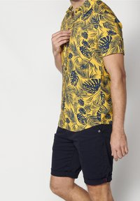 Homme portant une chemise jaune à manches courtes avec un motif de feuilles tropicales bleues et un short bleu marine foncé retroussé, debout devant un fond blanc.