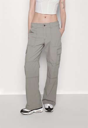 Person iført lysegrå cargobukser, hvide og sorte sneakers og en hvid cropped top, stående med hænderne i lommerne mod en ensfarvet baggrund.