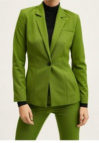 Femme portant un blazer ajusté vert citron et un pantalon assorti, sur un haut noir à col montant plissé, debout avec les mains détendues le long du corps.