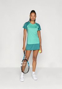 Tennisivorm teal ja tume teali värvuses, koos lühikeste varrukatega ülaosaga ja vastava seeliku ning mudel hoiab tennisereketit ja palli.