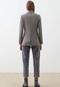 Šedé sako na míru s výraznými ramenními vycpávkami, rozparkem vzadu a zapínáním na knoflíky na manžetách, v kombinaci se šedými džínovými kalhotami a kotníkovými kozačkami s potiskem.