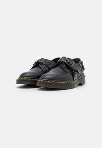 Scarpe in pelle nera con punta arrotondata, fascetta con fibbia e cuciture gialle lungo la suola. La suola in gomma testurizzata offre aderenza.