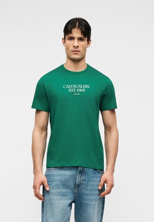 Uomo che indossa una t-shirt verde Calvin Klein con scritta bianca "Calvin Klein EST. 1968", abbinata a jeans azzurri, in piedi contro uno sfondo bianco semplice.