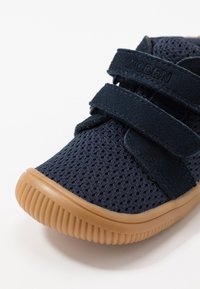 Námořnicky modrá síťová bota s Velcro pásky, gumovou podešví a strukturovaným povrchem. Disponuje zaoblenou špičkou a zesílenými okraji pro větší odolnost.