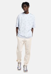 Ανοιχτό μπλε T-shirt με διακριτικό επαναλαμβανόμενο μοτίβο, συνδυασμένο με κρεμ φόρμα με τρεις κάθετες ρίγες και πολύχρωμα αθλητικά παπούτσια.