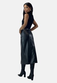 Falda de cuero sintético negra con corte recto, cintura alta y aberturas laterales, combinada con una blusa negra ajustada y botas de tacón altas negras hasta la rodilla.