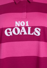 Rúžový a magentový pruhovaný polo tričko s golierom, s bielym vyšívaným textom "NO1 GOALS" vpredu. Mäkčký bavlnený materiál.