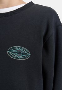 Svart hettegenser med ribbestrikket hals, med en oval logo i turkis og beige som sier "Billabong Boardshorts Since 1973." Myk stofftekstur.