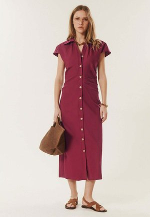 Femme portant une robe bordeaux mi-longue boutonnée, tenant un sac tressé marron, avec des sandales marron, des lunettes et un collier ainsi qu'un bracelet épais.