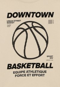 Bēša T-kreklu ar melnu tekstu un grafiku, tostarp basketbola dizainu un vārdiem "DOWNTOWN BASKETBALL" un "EQUIPE ATHLETIQUE."