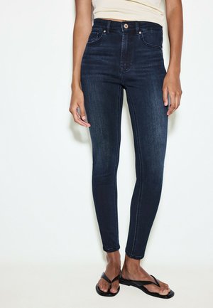 Person iført mørkeblå højtaljede skinny jeans og sorte flip-flops, stående mod en ensfarvet lys baggrund.