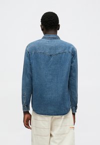 Chemise en denim bleu moyen avec manches longues, col pointu et détail de poche. Présente des poignets boutonnés et un ourlet arrondi à l'arrière.