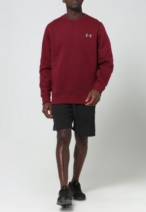 Sweatshirt bordeaux à manches longues et col rond, associé à un short noir et des chaussures de sport noires, avec un logo sous l'aisselle.