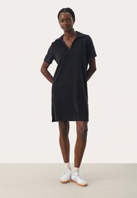 Abito polo nero realizzato in tessuto morbido, con colletto, maniche corte e spacchi laterali, abbinato a sneakers bianche.