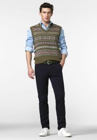Junger Mann trägt eine gemusterte olivgrüne Strickweste über einem hellblauen Hemd, dunkelblauen Hosen und weißen Sneakern, steht vor einem schlichten weißen Hintergrund.